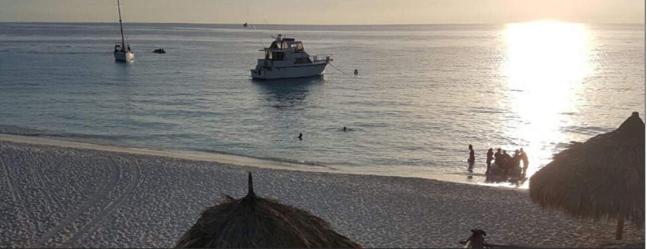Kamperen op Klein Curaçao biedt een serene zonsondergang op het strand met een kalme zee, kleine bootjes, een jacht voor de kust en mensen in silhouetten aan de rand van het water. Gezellige palapahutten, ideaal voor kamperen, staan verspreid over de zandkust.