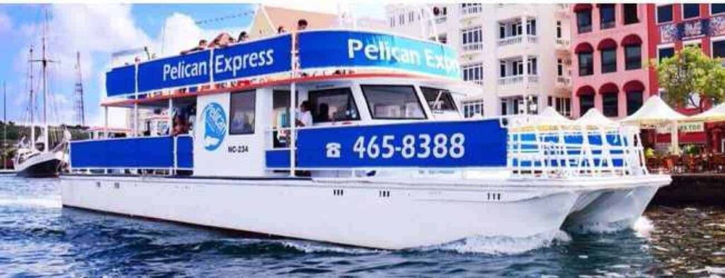 De wit met blauwe "Pelican Express" boot vaart langs de waterkant van een stad terwijl mensen genieten van het levendige uitzicht. De naam en het telefoonnummer worden weergegeven, terwijl gebouwen en tenten oplichten onder de Caribische zonsondergang.