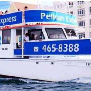 Caribbean sunset: The Pelican Express, a white and blue boat (465-8388), sails near a dock as passengers enjoy views of vibrant waterfront buildings and white tents under a partly cloudy sunset sky in the Caribbean.