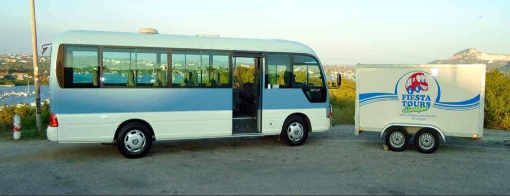 A light blue and white minibus with open doors is parked beside a FIESTA TOURS trailer, ready for the City and East Part Tour. Greenery, water, and distant hills appear in the background under a clear sky.