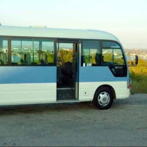 A light blue and white minibus with open doors is parked beside a FIESTA TOURS trailer, ready for the City and East Part Tour. Greenery, water, and distant hills appear in the background under a clear sky.