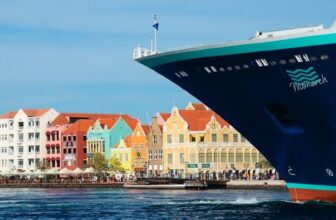 A large cruise ship’s bow dominates the right foreground near Curacao’s shoreline, lined with colorful Dutch-style buildings in pastel yellows, pinks, blues, and oranges, each topped with red-tiled roofs under a clear blue sky.