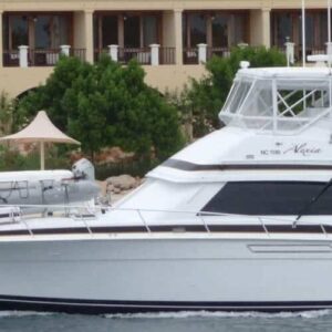 A white luxury yacht from Curacao boat tours and private charters cruises near a sandy shore with guests on board, as a resort with balconies, green trees, and beige sunshades overlooks the calm water.
