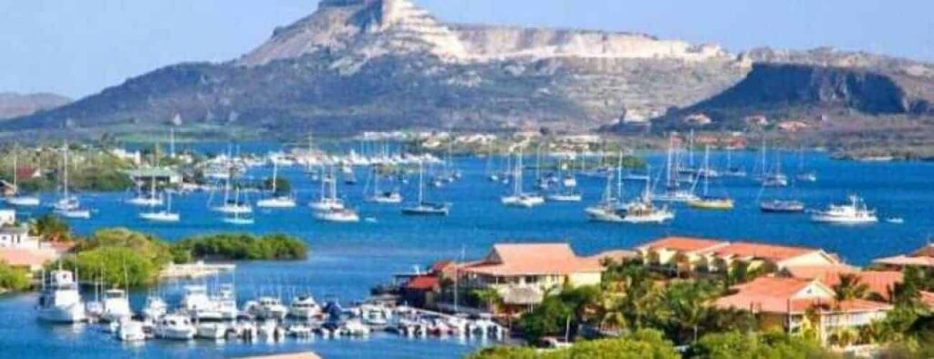 Curacao Land and Sunset features a colorful coastal scene with anchored boats in a blue bay, red-roofed houses, palm trees by the shore, and a glowing flat-topped mountain at sunset beneath a clear sky.