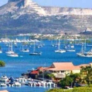 Curacao Land and Sunset features a colorful coastal scene with anchored boats in a blue bay, red-roofed houses, palm trees by the shore, and a glowing flat-topped mountain at sunset beneath a clear sky.