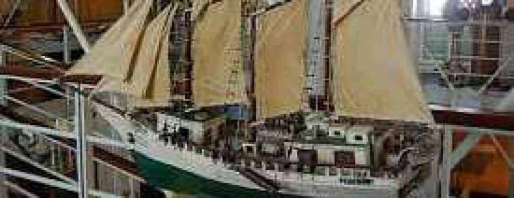 Het Maritiem Museum Curaçao heeft een gedetailleerd scheepsmodel met crèmekleurige zeilen en een groene romp, binnen tentoongesteld tussen witte balustrades en metalen constructies, die het rijke maritieme erfgoed van Curaçao laten zien.