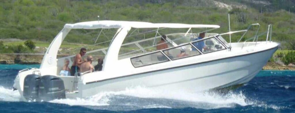 A white speedboat with a canopy speeds across blue water near Curacao, carrying several people enjoying their Curacao Speedboat Adventure and snorkeling along the lush, hilly coastline under a clear sky.