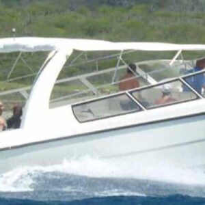 A white speedboat with a canopy speeds across blue water near Curacao, carrying several people enjoying their Curacao Speedboat Adventure and snorkeling along the lush, hilly coastline under a clear sky.