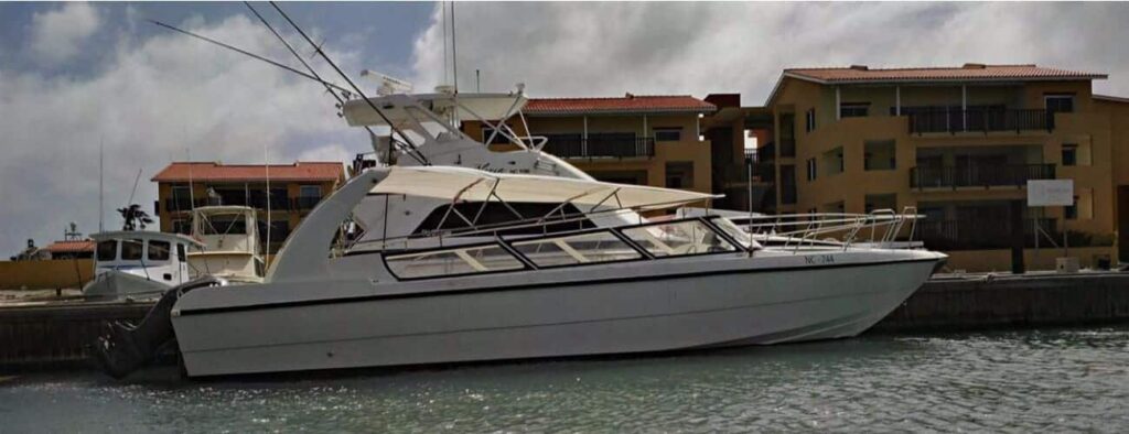 De Curaçaose watertaxi, een wit motorjacht met een luifel, vishengels en meerdere ramen, ligt aangemeerd in een jachthaven op Curaçao naast bruine gebouwen met rode daken. De weerspiegeling glinstert op het kalme water onder een gedeeltelijk bewolkte hemel.
