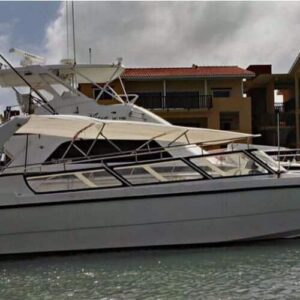 A white motor yacht with two fishing rods is docked by a concrete waterfront in Curacao. Yellow buildings with red-tiled roofs are in the background under a cloudy sky, and the Curacao watertaxi is partially visible on the left.