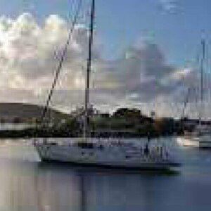 Experience a Day sail tour aboard white sailboats anchored in a marina at sunrise or sunset, with hills and a partly cloudy sky in the background. Gentle reflections of boats and clouds shimmer on the calm water’s surface.