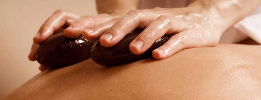 Close-up of two hands placing smooth, dark stones on a person's back during a Deep Tissue Massage, with shiny skin and stones suggesting warmth and relaxation in a spa setting.