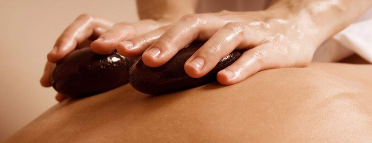Close-up of two hands placing smooth, dark stones on a person's back during a Deep Tissue Massage, with shiny skin and stones suggesting warmth and relaxation in a spa setting.