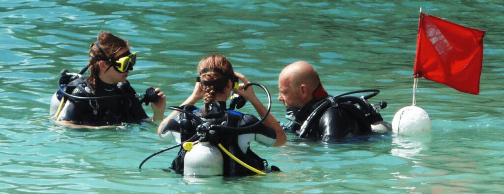 Three scuba divers in wetsuits stand in clear, shallow water during a Discover Scuba Diving or introduction dive. One adjusts their mask while the others watch. A red dive flag on a white buoy marks their adventure in calm turquoise waters.