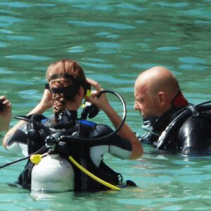 Three scuba divers in wetsuits stand in clear, shallow water during a Discover Scuba Diving or introduction dive. One adjusts their mask while the others watch. A red dive flag on a white buoy marks their adventure in calm turquoise waters.