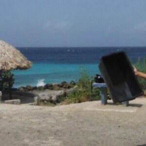 A man in sunglasses, a white tee, and patterned shorts poses with arms raised, holding "Dive in Playa Wachi" at the beach entrance, with thatched huts, turquoise ocean, and clear sky in the background.