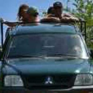 Three people enjoy Driving on a dirt path in a dark green SUV, surrounded by dense green trees and bushes under a clear blue sky. They sit atop the vehicle holding onto the roof rack as it moves forward.