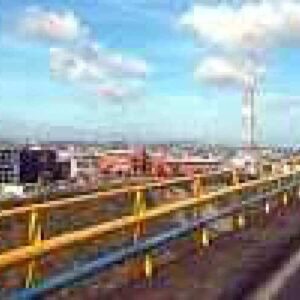 Op een heldere dag rijden twee mensen op een motor over een verhoogde East Side-snelweg met gele en blauwe vangrails. Industriële gebouwen en een gedeeltelijk bewolkte lucht zijn op de achtergrond te zien tijdens de East Side rondleiding.