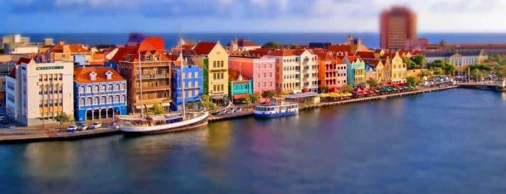 On the East to West Island Tour, brightly colored waterfront buildings and docked boats create a picturesque scene, their reflections shimmering in calm water beneath a partly cloudy sky with the sea in the distance.