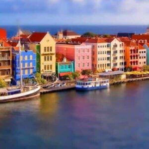 On the East to West Island Tour, brightly colored waterfront buildings and docked boats create a picturesque scene, their reflections shimmering in calm water beneath a partly cloudy sky with the sea in the distance.