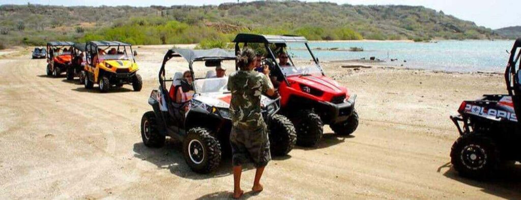 A person in shorts and a camo shirt stands on a sandy road beside four parked buggies during an exciting EAST-Tour by buggy, with a blue lake and green hills under a clear sky in the background.