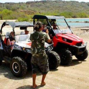 A person in shorts and a camo shirt stands on a sandy road beside four parked buggies during an exciting EAST-Tour by buggy, with a blue lake and green hills under a clear sky in the background.