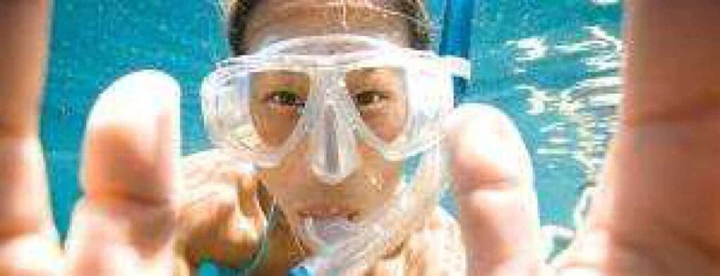 With "Everyone can snorkel!" swim goggles, a snorkeler faces the camera underwater with hands reaching forward; their facial features are visible through the clear goggles amid shimmering blue water and bubbles.