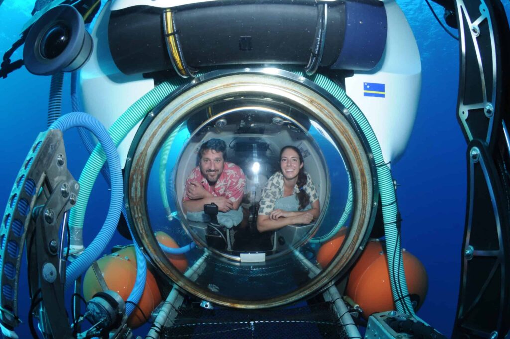 A man and woman smile and lean forward inside an Explorer dive submersible, visible through a spherical window with blue hoses, orange flotation devices, and deep blue ocean water surrounding them.