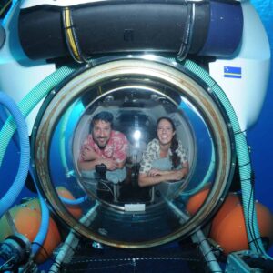 A man and woman smile and lean forward inside an Explorer dive submersible, visible through a spherical window with blue hoses, orange flotation devices, and deep blue ocean water surrounding them.
