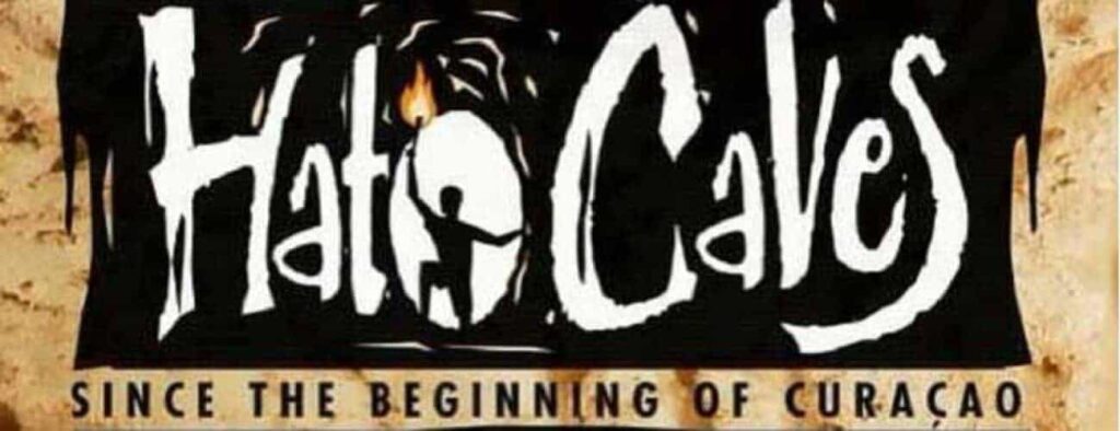 A bold white sign reads “Hato Caves,” with the “O” as a cave silhouette showing a person with a torch, promoting the Guided Cave Tour. Below, smaller text states “Since the beginning of Curaçao” against a textured brown rock background.