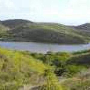 A tranquil lake bordered by green hills and shrubs under a partly cloudy sky creates an ideal spot for relaxation or as a starting point for your Guided Mountain Climb adventure in the nearby highlands.