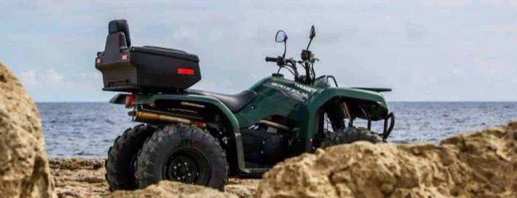 A dark green ATV from the Half Day East Side ATV Adventure, equipped with a rear storage box, is parked on sandy ground by large rocks, facing the calm sea under a partly cloudy sky. The ATV points away from the camera toward the water.