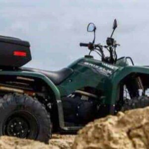 A dark green ATV from the Half Day East Side ATV Adventure, equipped with a rear storage box, is parked on sandy ground by large rocks, facing the calm sea under a partly cloudy sky. The ATV points away from the camera toward the water.