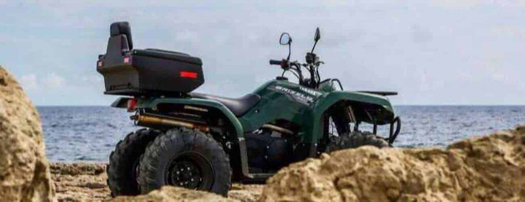 A Half day Westside ATV Adventure vehicle with large tires and a rear storage box sits on rocky shore facing the ocean, framed by tan rocks and a cloudy sky in this rugged coastal setting.