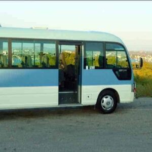 A blue and white tour bus with Fiesta Tours trailer, parked by green hills and water, awaits your Hato Caves and Ostrich Farm Tour adventure.