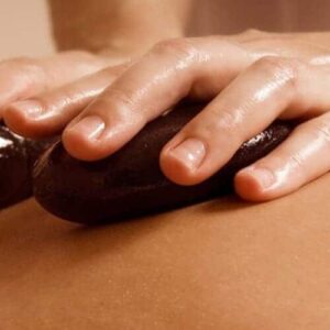 Close-up of hands pressing two smooth, dark stones onto a person's bare back during a Hotstone Massage, highlighting relaxation and wellness with a neutral, blurred background.