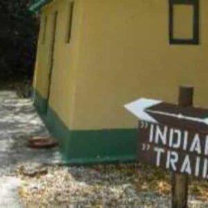 A dirt path runs past a small yellow and green building. In front, a wooden sign with white letters and arrows points right, marking the Indian Trail. Trees line the path, with dry leaves scattered on the ground. Product name: Indian Trail.
