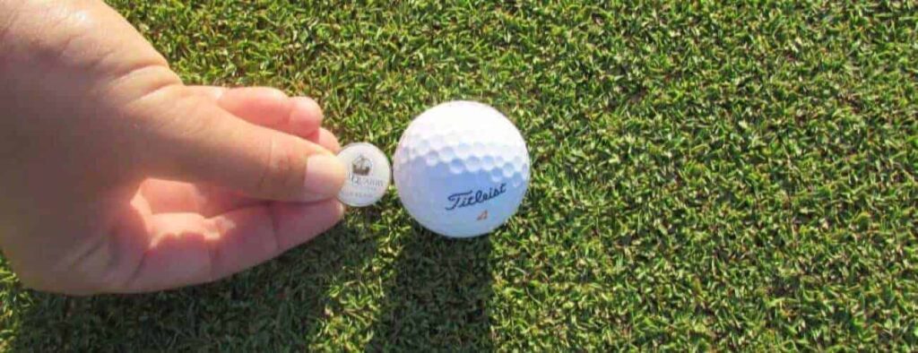 A hand places a round, flat marker displaying text and an image next to a white Titleist golf ball on green grass, possibly during an Intermediate 1 day Package training session on a golf course.