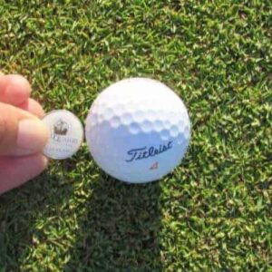A hand places a round, flat marker displaying text and an image next to a white Titleist golf ball on green grass, possibly during an Intermediate 1 day Package training session on a golf course.