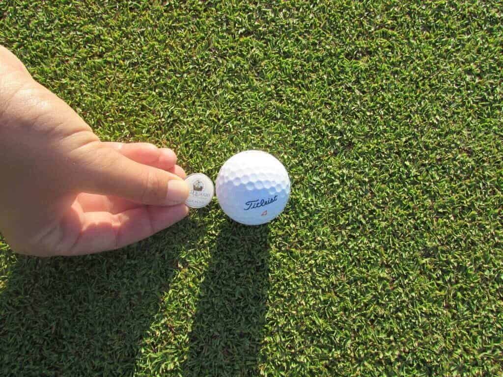 A hand places a round golf ball marker by a white Titleist ball on a lush green, bathed in sunlight—perfect for enjoying the Intermediate 2 day Package on this beautifully manicured course.