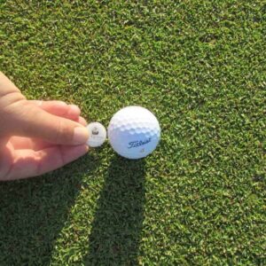 A hand places a round golf ball marker by a white Titleist ball on a lush green, bathed in sunlight—perfect for enjoying the Intermediate 2 day Package on this beautifully manicured course.