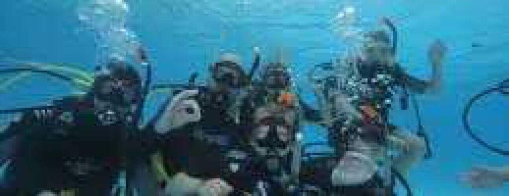 Five people in scuba gear are underwater in a clear blue swimming pool, participating in an Intro Dive. Wearing masks and tanks, they sit or kneel on the pool floor with bubbles rising. One person on the right waves at the camera.