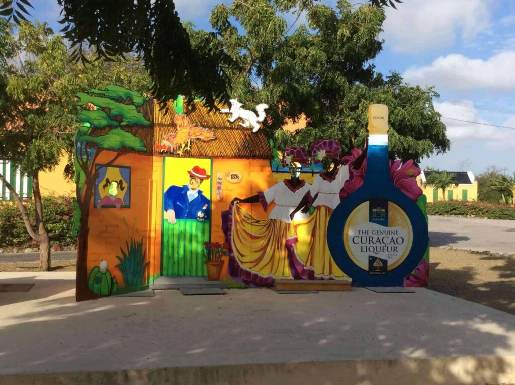 A vibrant cutout display for the Island and Beach Tour features two women in yellow skirts dancing, a man in blue by a green door, tropical plants, and colorful buildings—capturing the lively spirit of an island adventure.