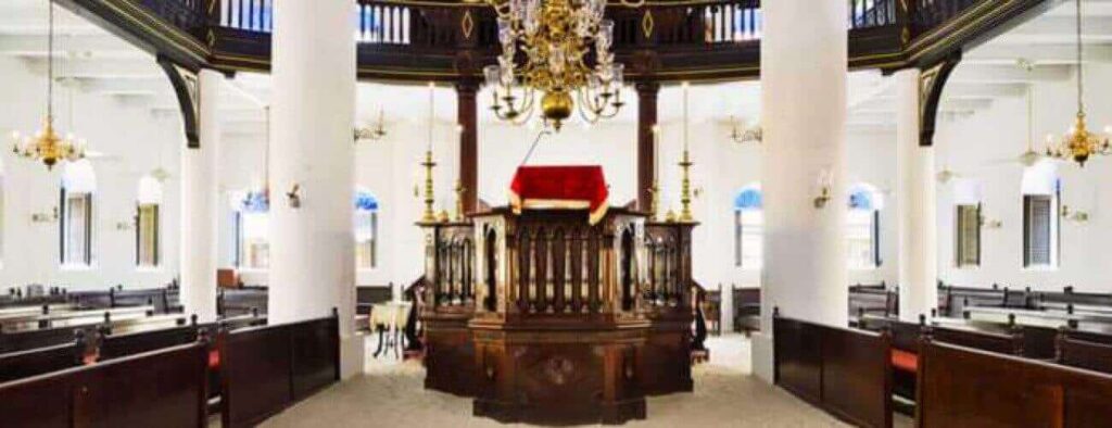 The Jewish Heritage Tour features the interior of a historic synagogue with white walls, tall columns, dark wooden pews, a red-draped bimah, gold chandeliers, and sunlight streaming through arched windows—a true tour highlight.