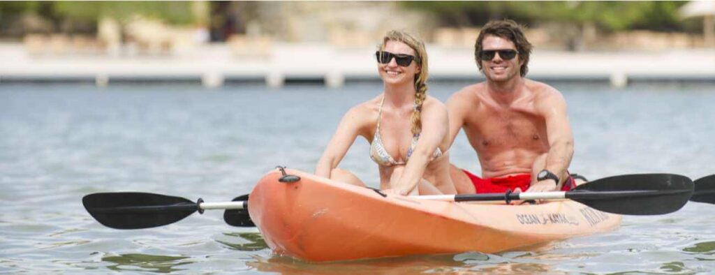 A smiling woman in a bikini and a shirtless man, both wearing sunglasses, paddle an orange tandem kayak on calm water during the Kayak Snorkel Tour Cas Abao - Porto Marie or Daaibooi (2.5hr), with greenery and shoreline in the background.