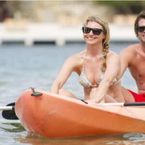 A smiling woman in a bikini and a shirtless man, both wearing sunglasses, paddle an orange tandem kayak on calm water during the Kayak Snorkel Tour Cas Abao - Porto Marie or Daaibooi (2.5hr), with greenery and shoreline in the background.