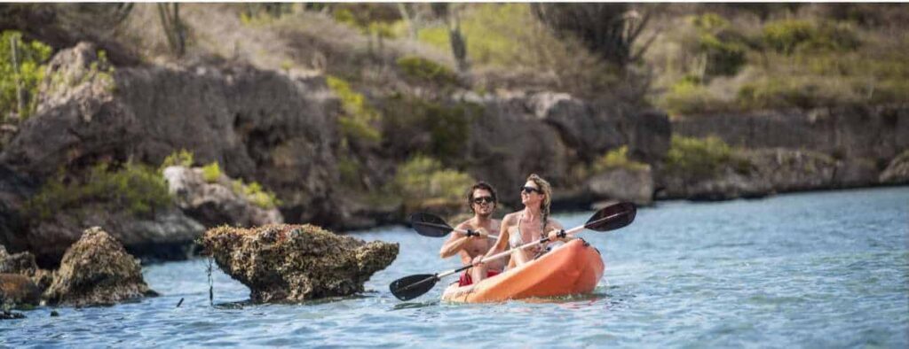 Two people wearing sunglasses paddle a bright orange kayak on clear blue water near Cas Abao’s rocky shores and lush greenery, enjoying sunny weather during their Kayak Snorkel Tour Cas Abao - Sunset Waters (4hr).