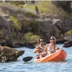 Two people wearing sunglasses paddle a bright orange kayak on clear blue water near Cas Abao’s rocky shores and lush greenery, enjoying sunny weather during their Kayak Snorkel Tour Cas Abao - Sunset Waters (4hr).