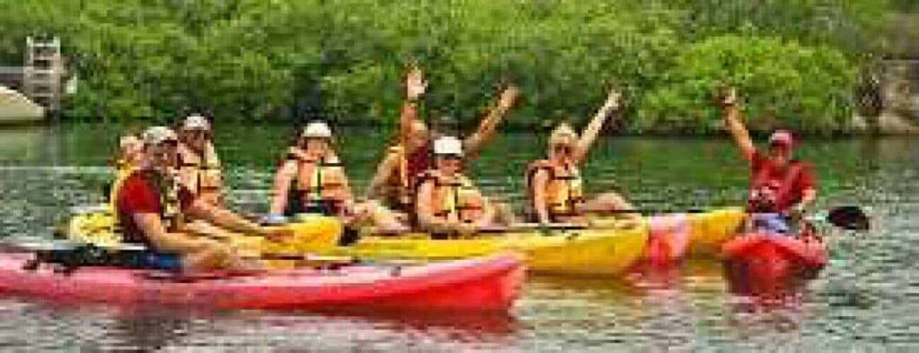 A group in life jackets and helmets enjoy "Kayaking on the Spanish Water," smiling and raising their arms in red and yellow kayaks as they float on calm waters, surrounded by green shrubs and trees for a cheerful outdoor adventure.