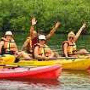 Een groep mensen geniet van Kajakken op het Spaanse Water in kleurrijke kajaks, omringd door groen gebladerte. Ze dragen zwemvesten en hoeden, peddelen samen en glimlachen, waardoor er een vrolijke en levendige sfeer op het kalme water ontstaat.